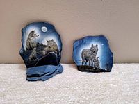 Two slate sculptures featuring wolf themes, one with three wolves on a blue base and the other with a standing wolf and pup on slate without base.