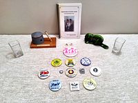 Top-down photo showing the full lot on a white textured surface: spiral-bound recipe book, Murney Tower pen holder with gold pen, green beaver sculpture, two clear shot glasses, and a group of 14 souvenir pins with various Kingston logos and text.