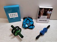 Photo showing Body Rock 8 lb weighted vest packaging, vest folded, ab wheel roller, and double-ball massage roller on floor.
