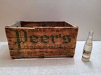Side view of vintage wooden Peer's soda bottle crate with green branding and a Peer’s soda bottle placed next to it.