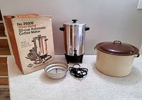 Photo of the vintage West Bend 30-cup automatic coffee maker with its box, filter insert, power cord and the large enamel pot with lid on a table.