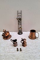 Miniature vintage brass and copper items on white surface including ladder with two figures, oil lamp, tankard, pitchers, and candlesticks.