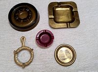 Five vintage ashtrays arranged on a light fabric surface, showing mix of brass and glass materials, various shapes and colors