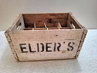 Angled front view showing the 'ELDER'S' black stencil on white painted wood crate and cutout handles on each side.