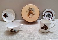 Five bone china items with horse head designs: a mustache cup and plate set, Wellington mug, and three ashtrays, displayed along with a round copper tin.