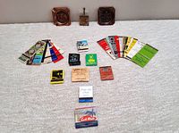 Assortment of colorful vintage matchbooks and covers laid out on a light surface with three mini copper ashtrays in the background.