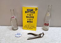 Five collectible items displayed against a neutral background on white fabric: two vintage soda bottles, a yellow collectors price guide book, a metal flip top bottle opener, and a round pinback button