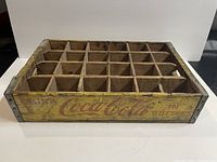 Photo showing front side of wooden crate with yellow paint and faded red Coca Cola logo and "IN BOTTLES" text, featuring 24 bottle compartments.
