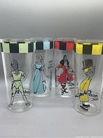 Four tall cylindrical drinking glasses with colored checkered rim bands, each featuring different Peter Pan characters in color