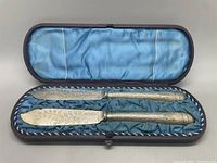 Pair of antique butter knives with ornate engraved blades and detailed textured handles, displayed inside original hinged box with blue silk interior lining.