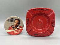 Red metal Coca Cola square ashtray with text and logo, alongside a round promotional button featuring Bill Crosby and vintage slogans.