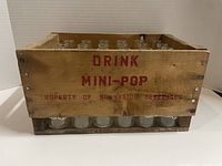Wooden crate showing front side with red text 'Drink Mini-Pop' and 'Property of Sunnyside Beverages'. Empty, clean clear glass bottles visible inside crate arranged in rows.