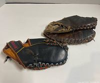 Photo showing two vintage early leather baseball gloves, one black and tan, the other dark brown, displayed flat to show exterior side.