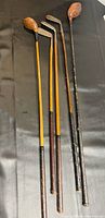 Photo showing five antique wooden golf clubs arranged on black surface, with details of shafts and heads visible to show condition and size.
