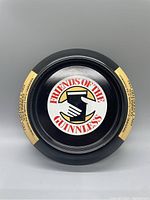 Front view of black circular ashtray with central logo showing two hands holding a pint and 'Friends Of The Guinness' text. Yellow printed side labels on rim.
