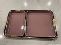 Set of three pink nesting trays with metal rims and wicker handles laid out on a white marble surface.