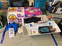 Photo showing the Six Million Dollar Man doll lying on the open Bionic Transport and Repair Station with the original box behind. Includes additional small items and accessories.