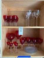 Wooden shelf with red and clear crystal glasses and clear glass plates. Red glasses have twisted stems, different shapes including small cocktail glasses and large goblets. Clear glasses are clear with elongated bowls and twisted stems. Plates are clear and stacked.