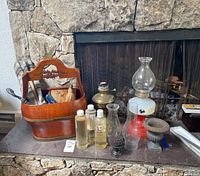 View of oil lamps, bottles of lamp oil, wooden caddy with fireplace tools, and decorative metal urn arranged on stone fireplace hearth.