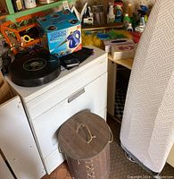 View of white clothes dryer with various cleaning supplies on top and beside it, laundry hamper in front, ironing board at side.