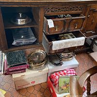 View of wooden cabinet with open drawers showing silverware and napkin rings, silver serving dishes on boxes beneath and checkered linens in box.