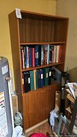 Full view of teak wood shelving unit showing three open shelves, two filled with books and binders, and a lower cabinet with sliding doors.