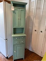Tall green wooden cabinet next to refrigerator, showing upper and lower cabinet sections and white knobs.