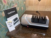 ChefsChoice electric sharpener with user manual on countertop with floral wallpaper background.