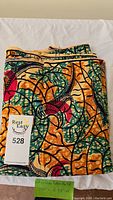 Folded colorful African batik cotton fabric with wavy, leafy abstract pattern in green, orange, red and black, folded on a white background and partially covered by a lot tag.