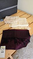 Three scarves on wooden table: two light cream-colored scarves with fringed edges and one dark purple scarf with black trim fringes and pattern.