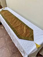 Full view of the brown table runner laid out on a white surface showing its length and styling with green borders and tassels.
