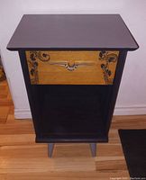 Front view of refinished vintage nightstand showing slate grey and matte black finish, original wood drawer with art deco floral detail and longhorn metal handle, angled legs.