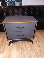 Front view of the nightstand showing its matte slate grey face, wood top and curved black base with two drawers closed and decorative handles.