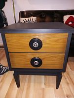 Front view of the nightstand showing matte black frame, two natural wood drawers, and round black knobs with metal decoration in center.