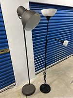 Two floor lamps, one industrial style with a tall cylindrical base and grey spotlight, the other black lamp with white upward shade and small reading lamp.