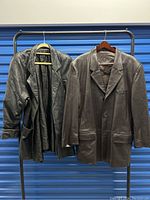 Two men's vintage leather jackets hanging on hangers, one black oversized size XS and one dark brown size 48R.