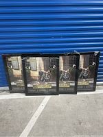 Four black poster frames leaning against blue storage door, some plastic wrapping partially removed, visible size label 22x28 inches
