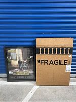 Single black poster frame displayed alongside a cardboard box labeled 'FRAGILE' indicating original packaging. The frame is 22 by 28 inches from the brand Studio Décor with a picture inside showing an outdoor scene with a bicycle.