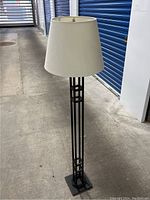 Full view of the floor lamp with black metal base and white lampshade, standing on concrete floor indoors.