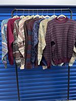 Eight vintage men's sweaters hanging on a rack. Various patterns and colors including blue, burgundy, beige, and mixed tones.