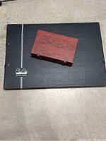 Black rectangular puzzle caddy with silver stripe and handle, closed, flat on a concrete floor with a smaller wooden box on top.