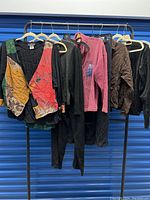 Full rack view showing 10 pieces of women's assorted art-to-wear outfits. Various jackets, sweaters, skirts, and pants hanging on rack with a blue background.