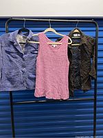 Three hanging garments including a blue textured jacket, pink sleeveless knit top, and dark brown eyelet-patterned jacket on a clothing rack.