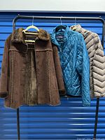 Three women's outerwear jackets on hangers against blue background: brown faux fur coat, blue quilted jacket, beige puffer jacket