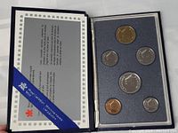 Photo of open blue presentation binder displaying six Canadian coins securely held in foam cutouts alongside certificate document.