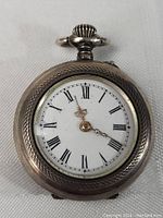 Front view of antique pocket watch showing white dial with black Roman numerals and ornate hands. Textured metal case and crown with loop at top visible.