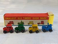 Four tiny antique metal toy cars in red, green, yellow, and blue arranged in front of original packaging box with vintage vehicle illustrations and 'Smallest Old Timers Ever Made' text.