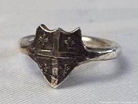 Front and side view of an early sterling silver ring with a shield-shaped face divided by a cross with fleur de lis in each quadrant, showing surface wear and tarnish.