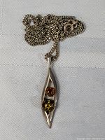 Close-up of pendant necklace lying on cloth showing two amber stones, orange and greenish-yellow, within silver elongated pendant and chain with clasp.