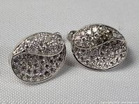 Pair of sterling silver earrings encrusted with numerous small clear stones captured against a plain background highlighting the front of both earrings.